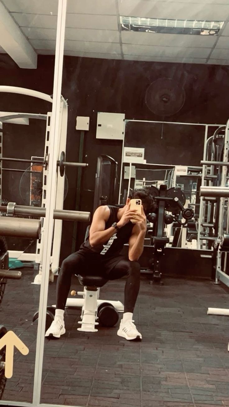 a man squats in the middle of a gym while taking a photo with his cell phone