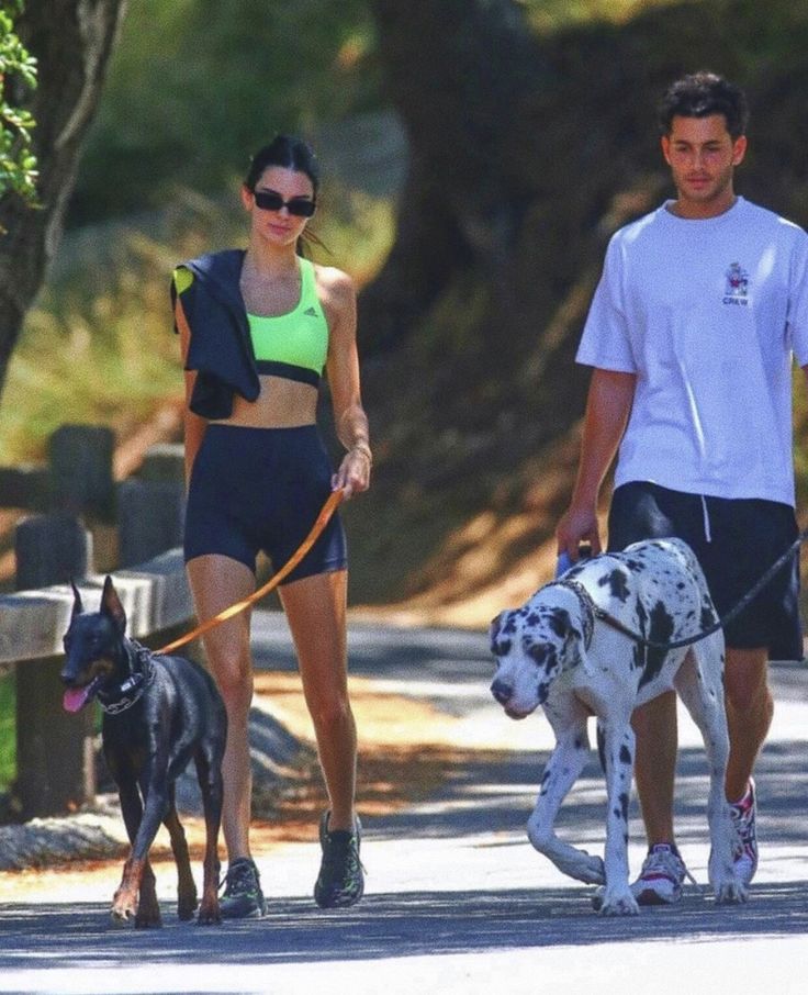 a man and woman walking two dogs on a leash down a street with trees in the background