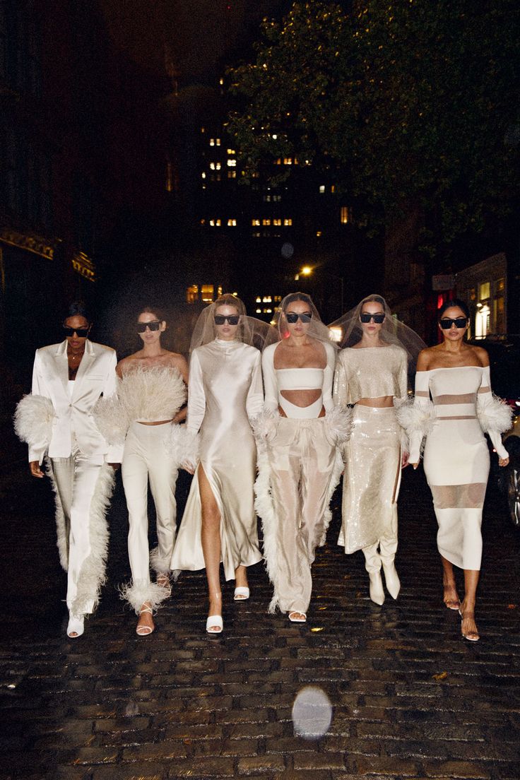 models in white outfits walk down the street at night with one woman wearing sunglasses on her head