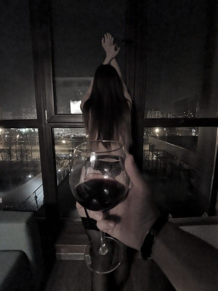 a person holding a glass of wine in front of a window with the city lights behind them