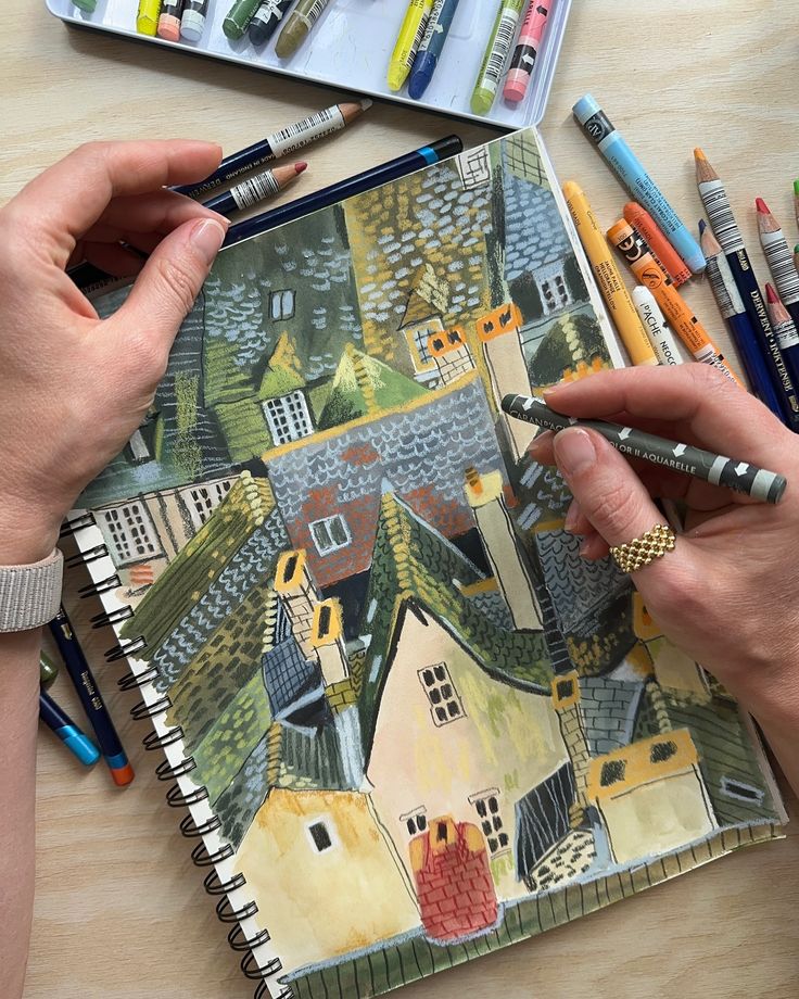 two hands are holding colored pencils over a drawing book with houses and buildings on it