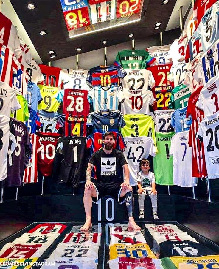 a man sitting on top of a table in front of many different colored shirts hanging from the ceiling