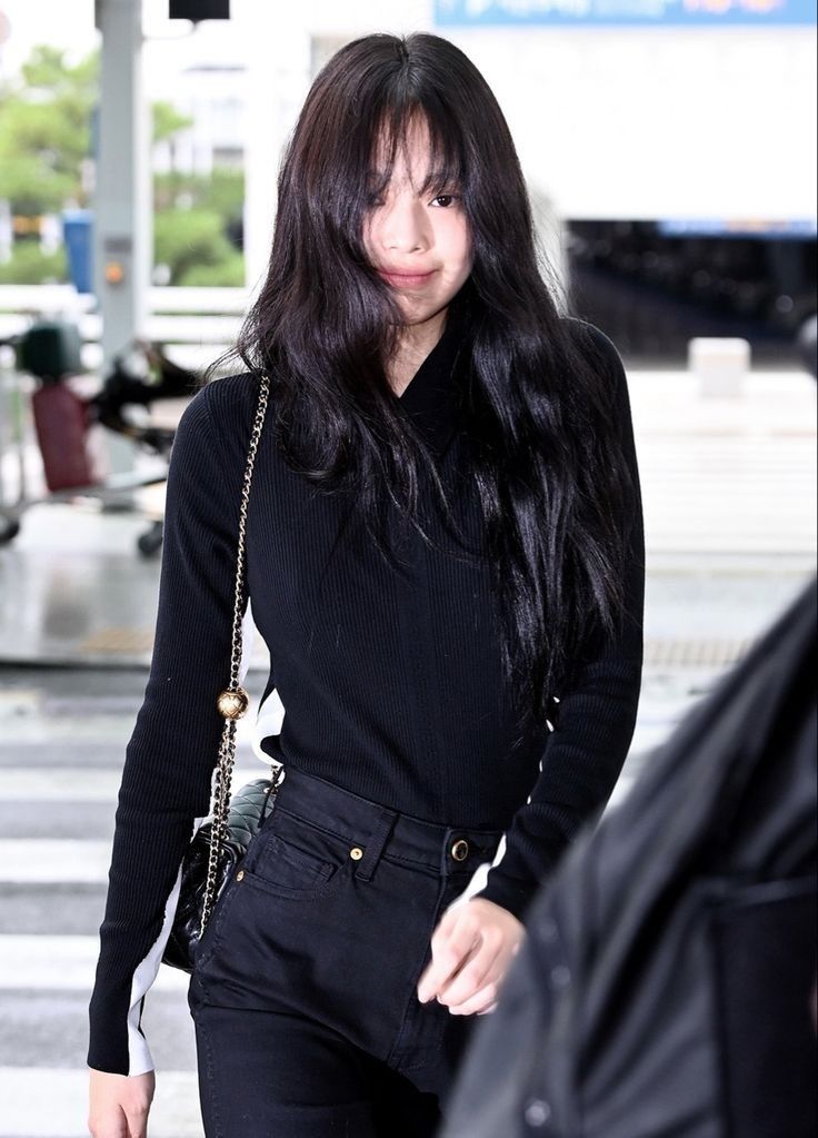 a woman with long black hair walking through an airport carrying a handbag and looking at the camera