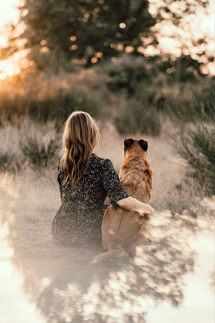 a woman sitting on the ground with her dog looking out at the trees in the distance