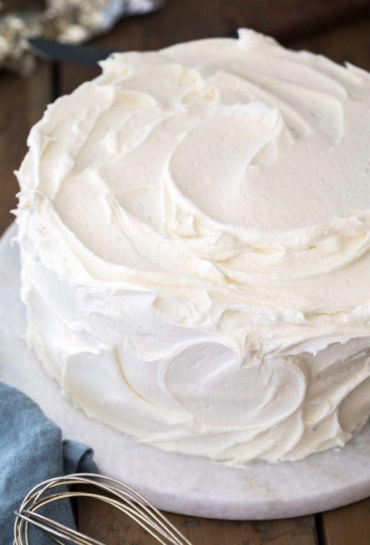 a cake with white frosting sitting on top of a table next to a whisk