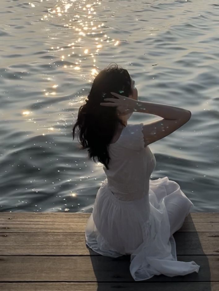 a woman sitting on a dock in front of the water with her hands behind her head