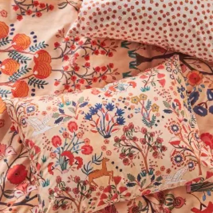 an assortment of pillows and blankets on a bed with floral designs in red, orange, yellow and blue