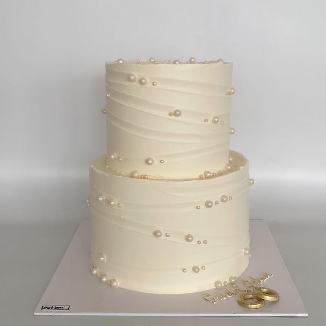 a three tiered white cake with pearls on the side and two gold wedding rings next to it