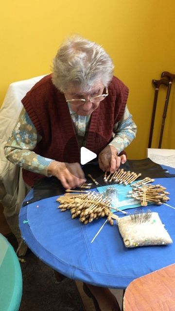 an older woman sitting at a table working on something with sticks and needles in front of her