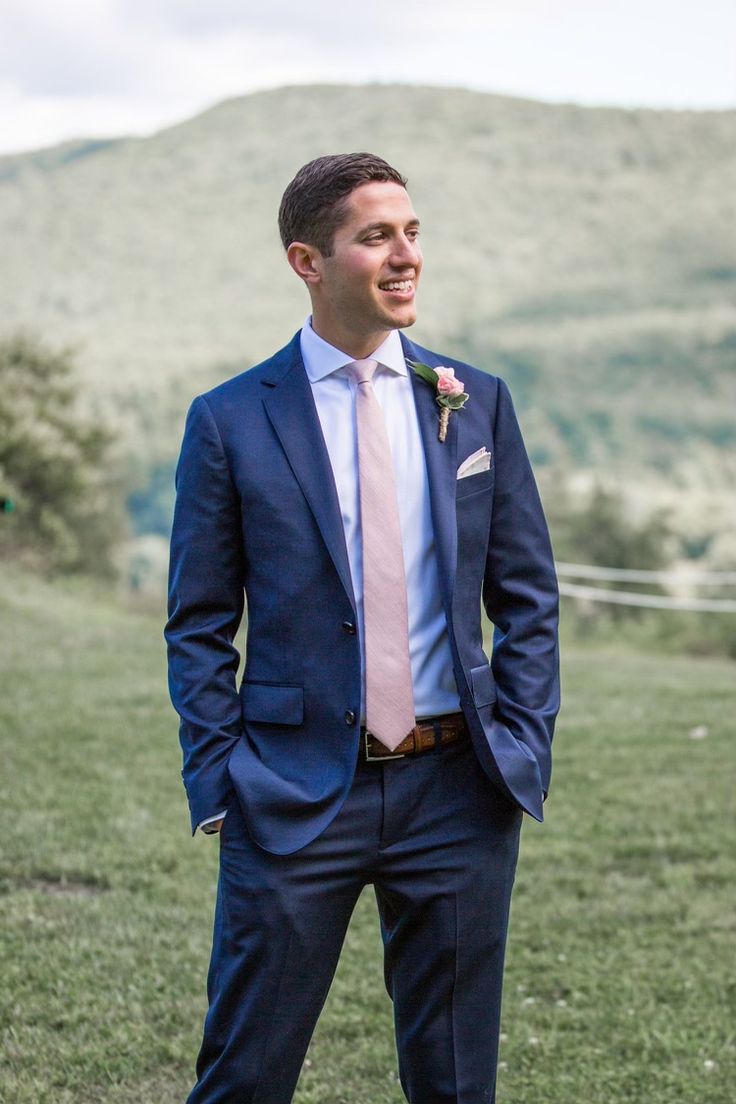 a man in a blue suit and pink tie is standing on the grass with his hands in his pockets