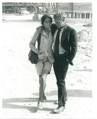 black and white photograph of man and woman walking in the sand with construction behind them