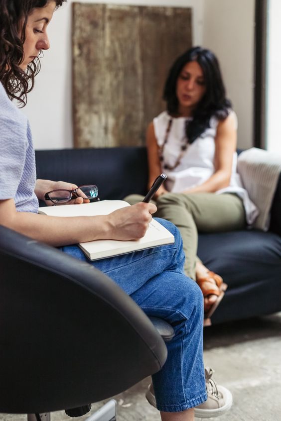 two women are sitting on a couch and one is writing in a notebook while the other looks at her phone