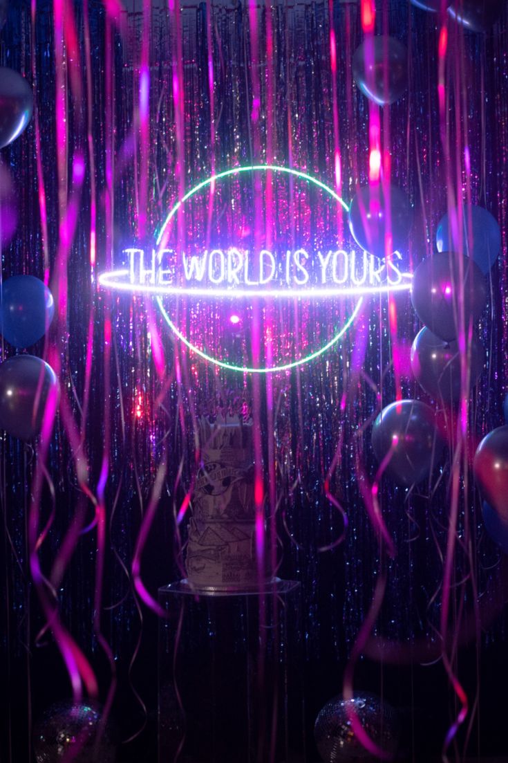 the world is yours neon sign surrounded by balloons and streamers in a dark room