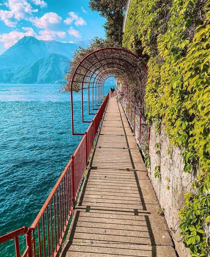 the walkway is lined with vines and railings along the water's edge, leading to mountains