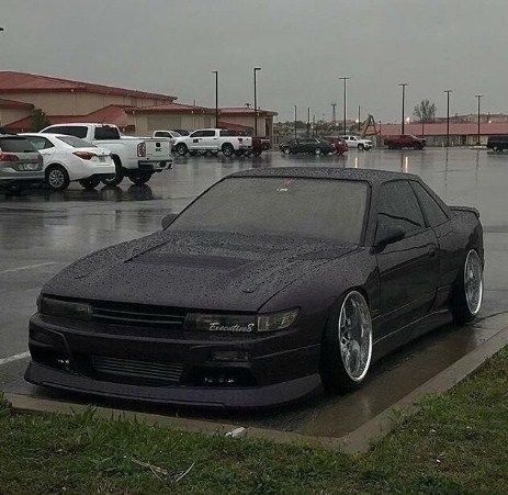 a black car parked in a parking lot on a rainy day with other cars behind it