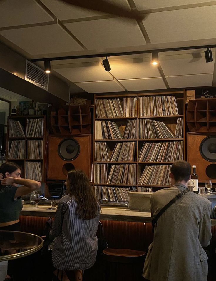 two people standing in front of a bar with records on the wall and shelves behind them