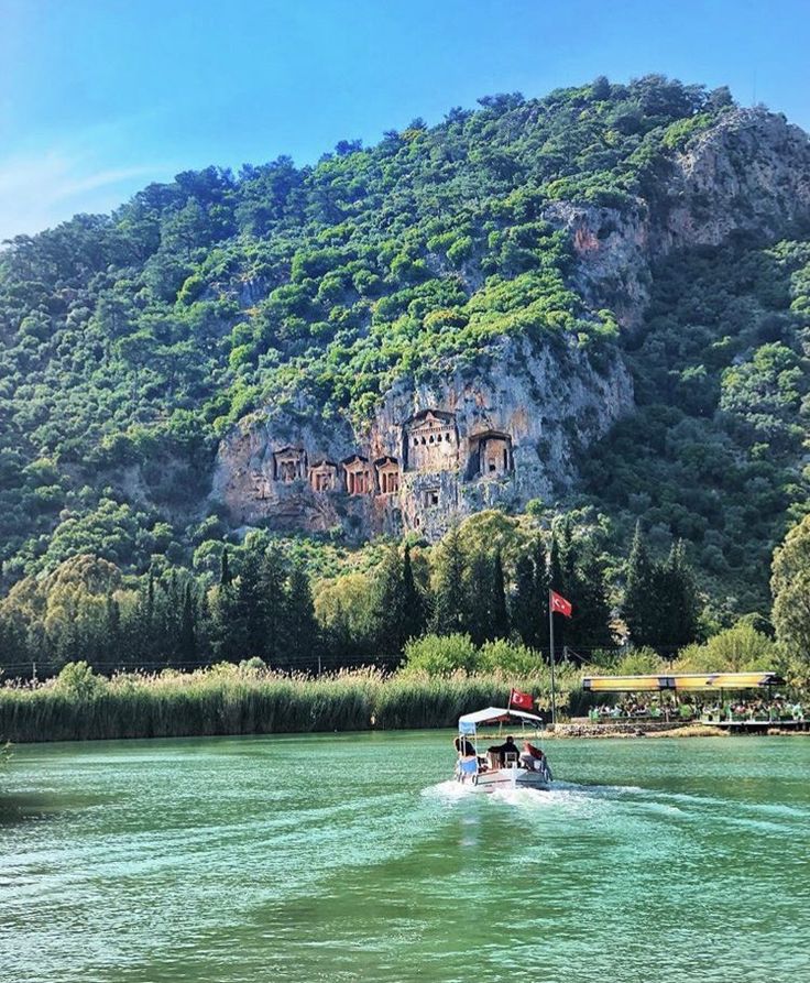 a boat traveling down a river next to a lush green forest covered mountain side under a blue sky