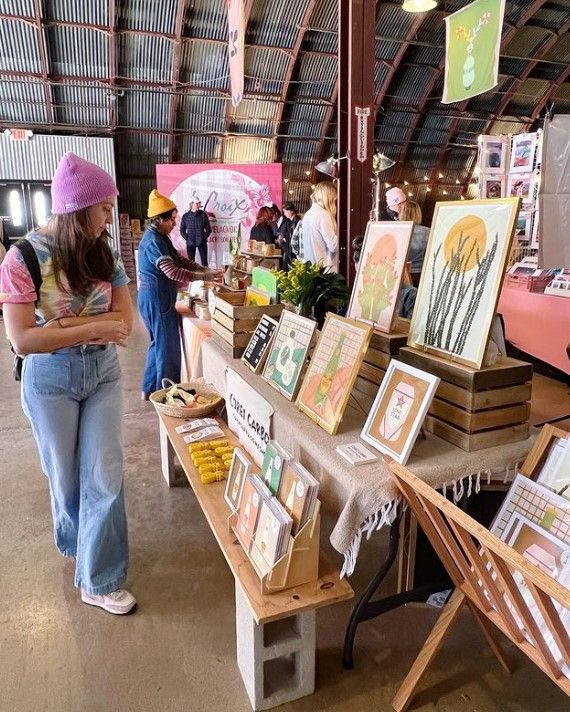 a woman standing next to a table with paintings on it and people looking at them