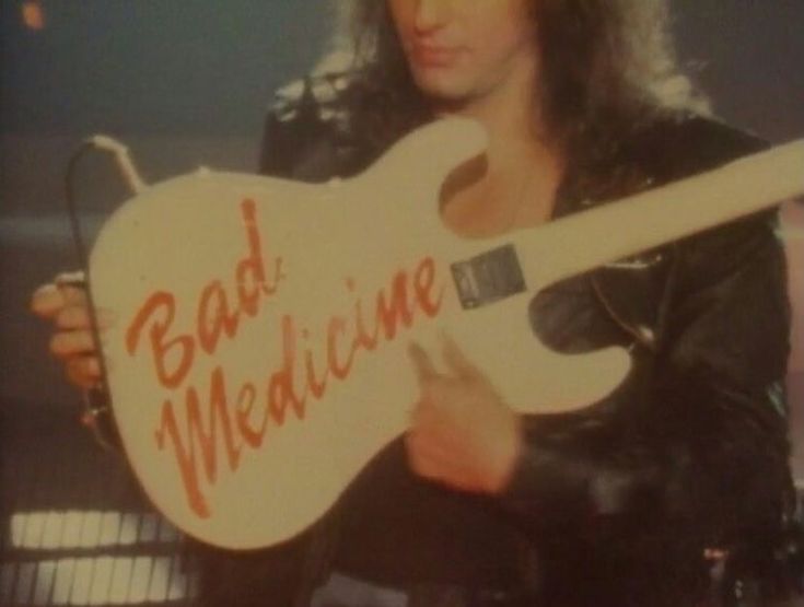 a man holding a white guitar with the words bad medicine written on it in front of him