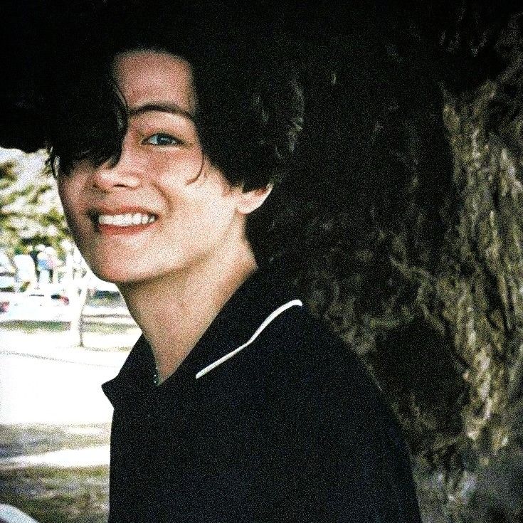 a young man smiling under a tree in the dark with his hair pulled back and wearing a black polo shirt