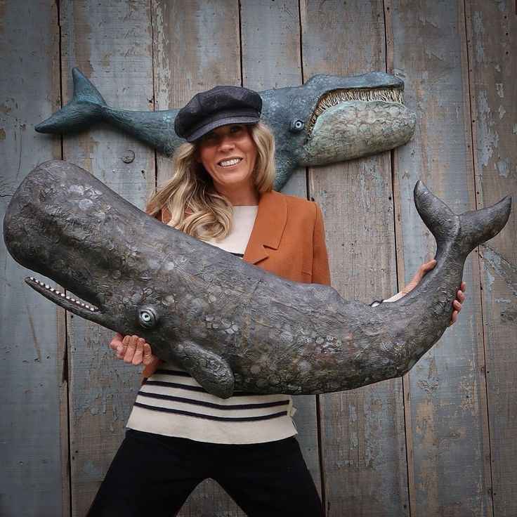 a woman is posing with a fake whale in front of a wooden wall and wearing a hat