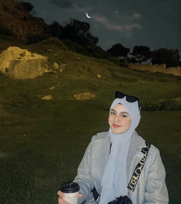 a woman wearing a hijab and holding a coffee cup in her hand while sitting on the grass