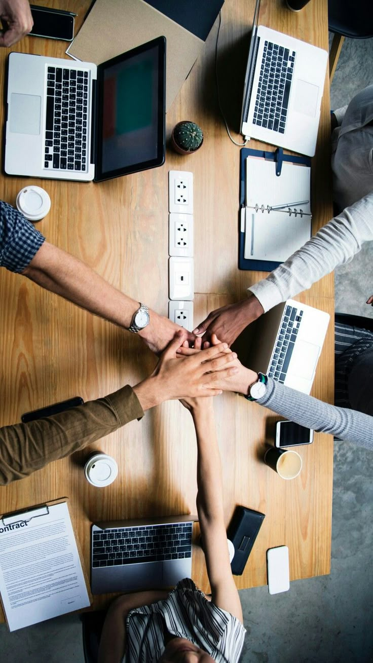 four people holding hands over a table with laptops