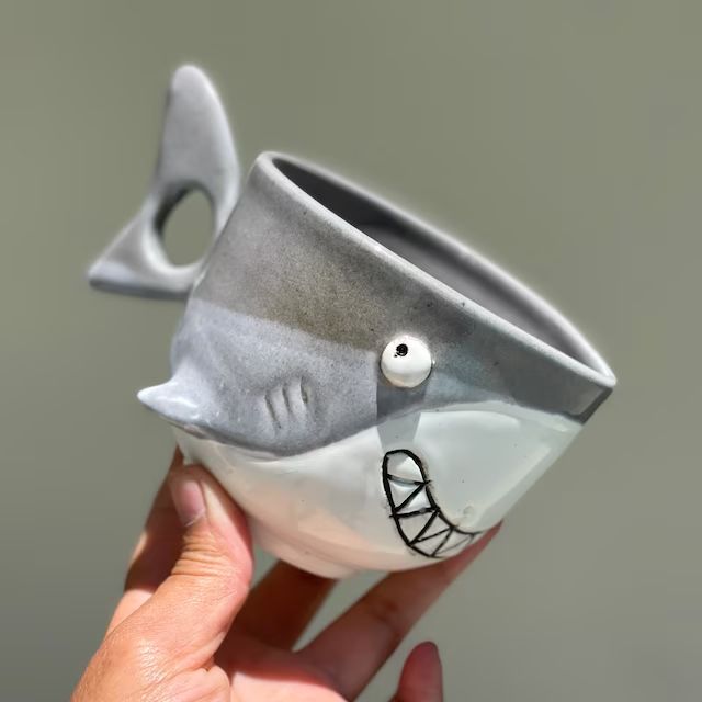 a hand holding a ceramic cup with a shark face on it