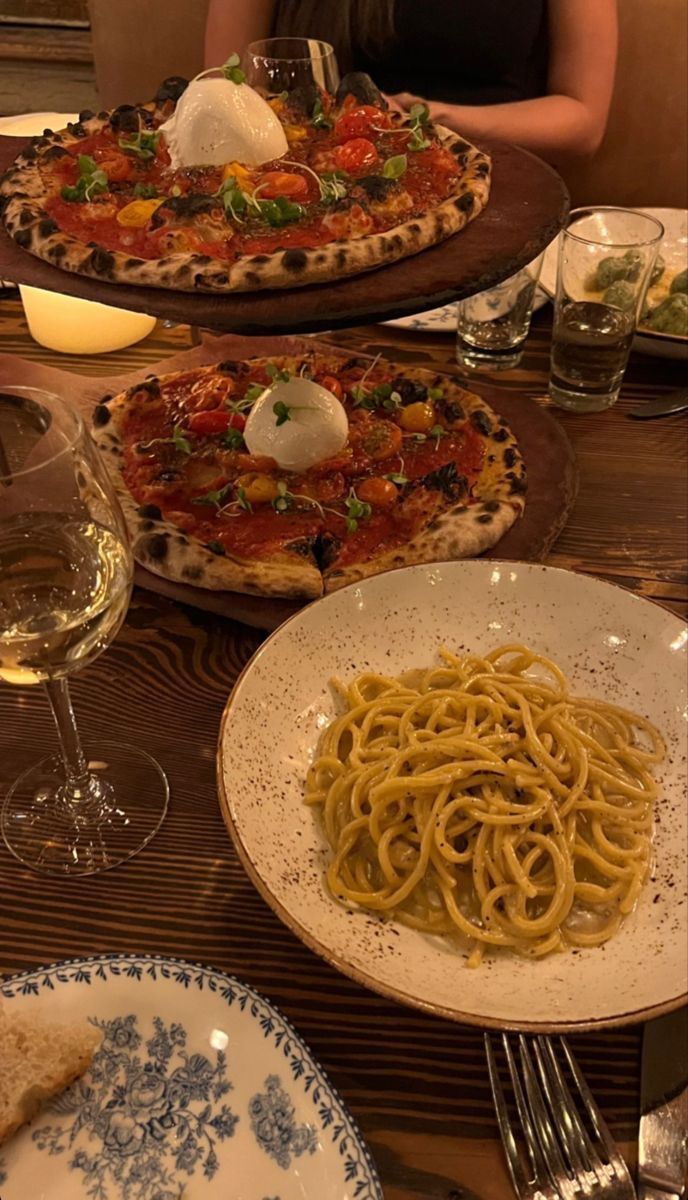 two pizzas and some pasta are on the table with wine glasses, silverware and candles