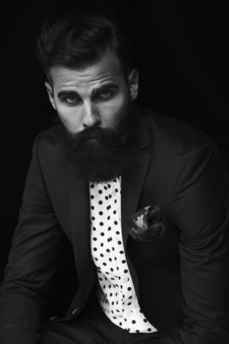 a man with a long beard wearing a suit and white polka dot tie sitting in front of a black background