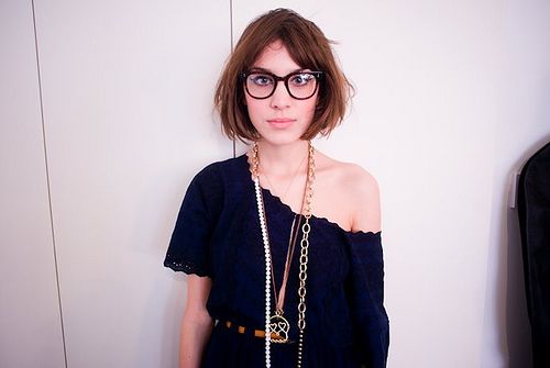 a woman wearing glasses standing in front of a wall with a chain around her neck