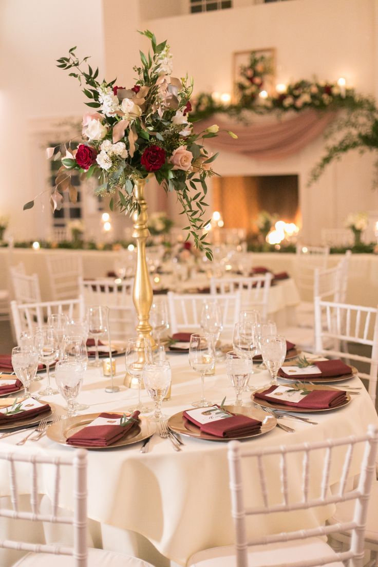 the table is set with white linens and red napkins, gold vase filled with flowers