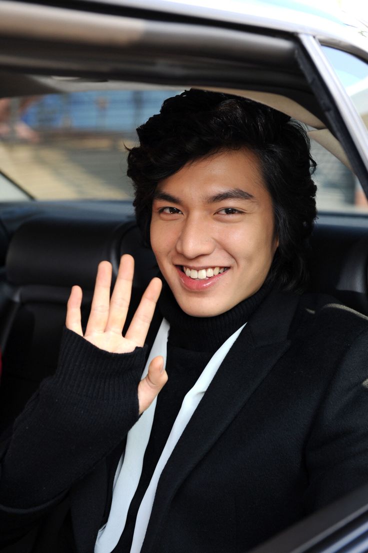 a man sitting in a car waving at the camera with his hand up and smiling