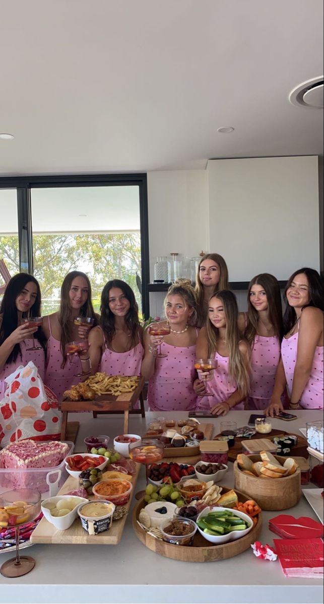 a group of women in pink dresses standing around a table full of food and drinks