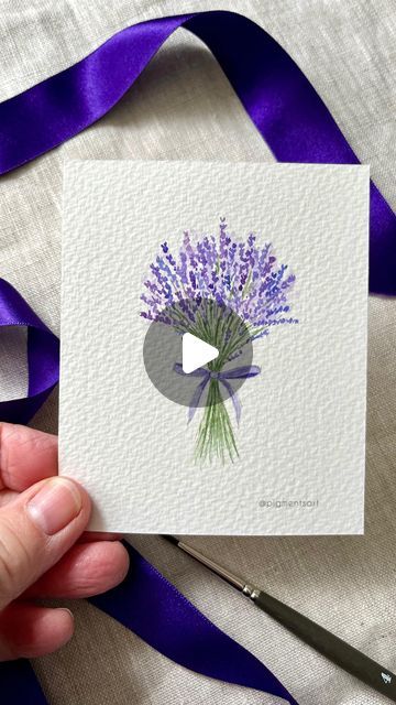 a hand holding up a card with purple flowers on it and a ribbon around it