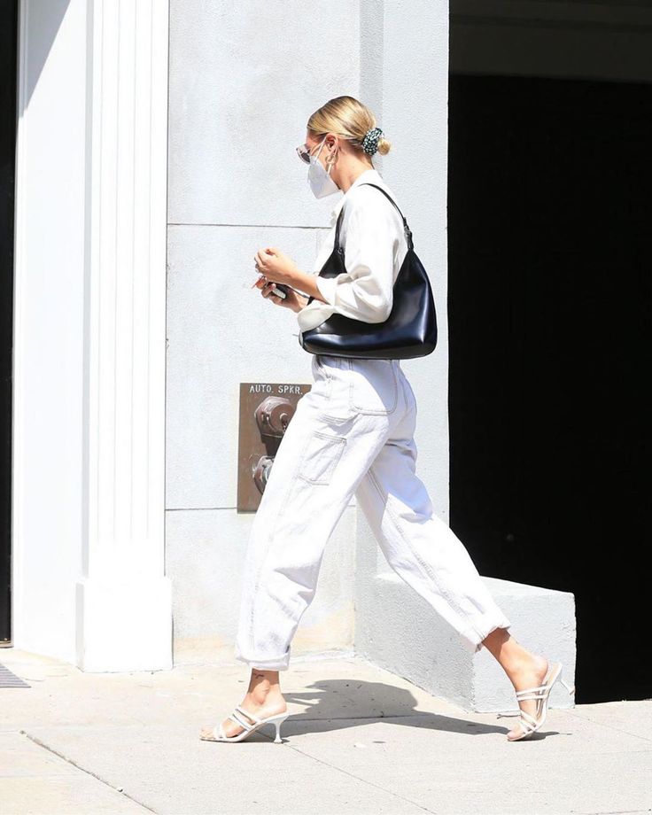 a woman walking down the street in white pants and heels, wearing a face mask