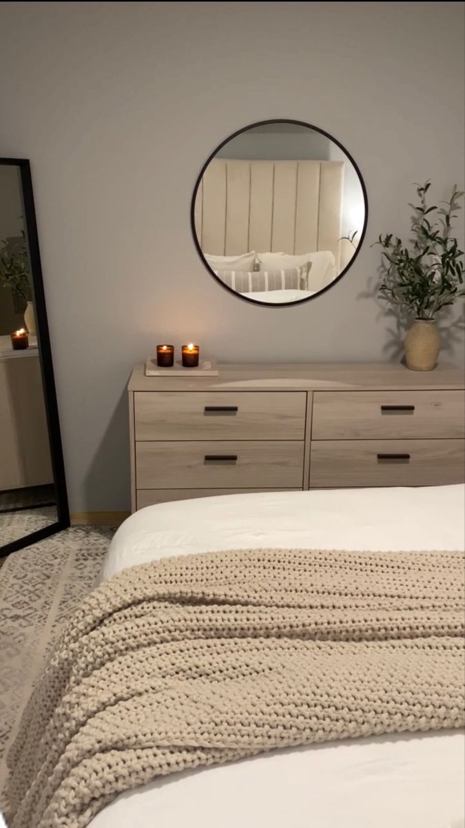 a white bed sitting under a round mirror next to a dresser with candles on it