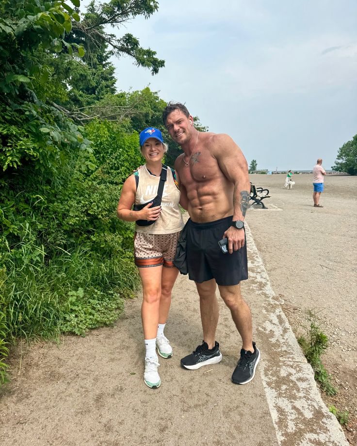 a man and woman standing next to each other on a path near the beach with trees