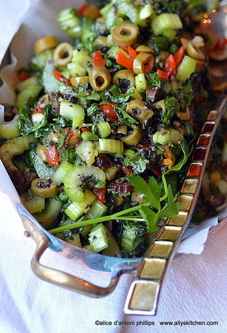 a salad with olives, peppers and other vegetables in a silver serving dish on a white table cloth