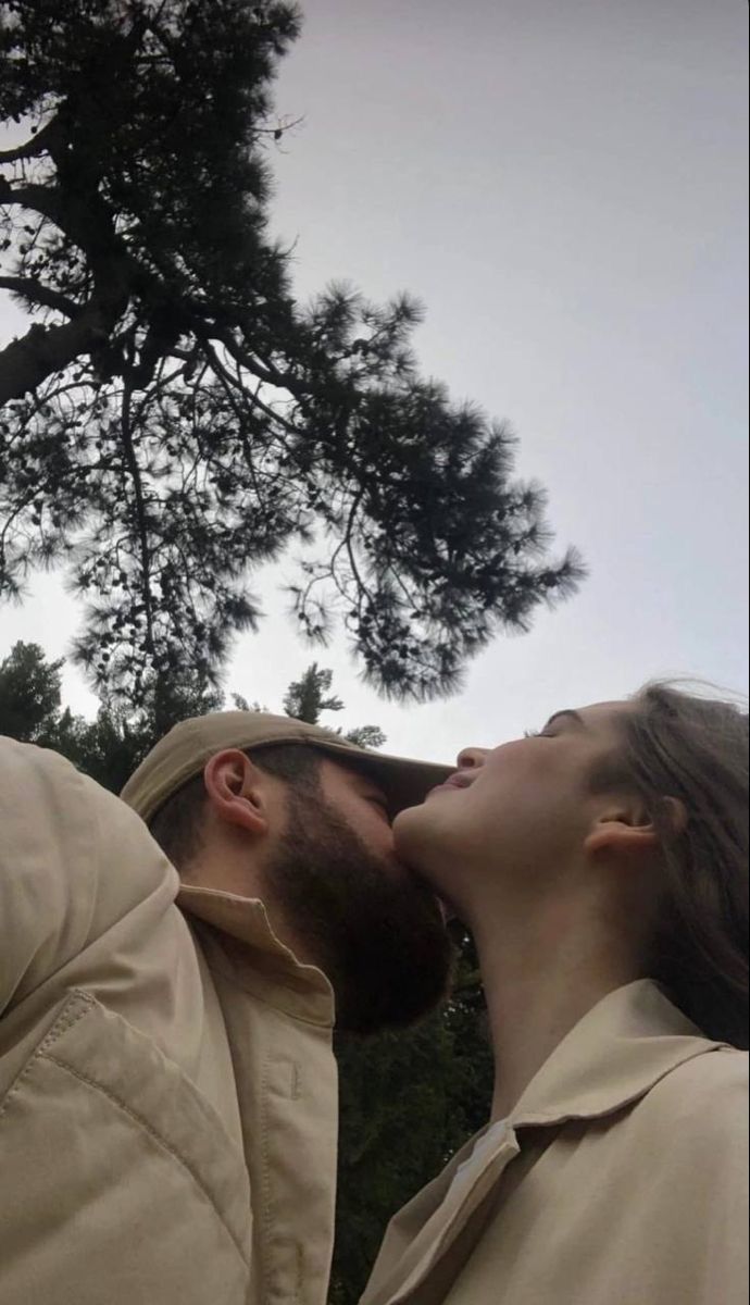 two people standing next to each other with trees in the background and one person kissing the other