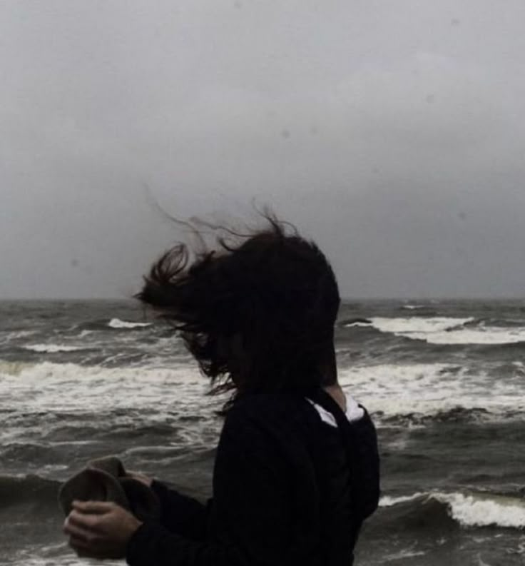 a woman standing on the beach with her hair blowing in the wind and looking at the ocean