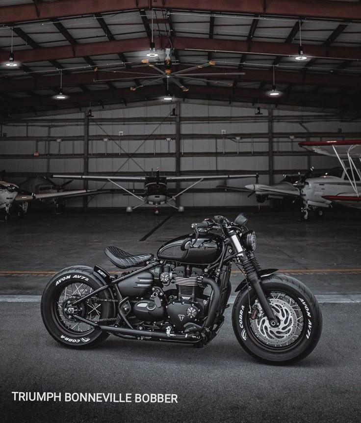 a black motorcycle parked in an airplane hanger with the caption triumph bonnetville bobber