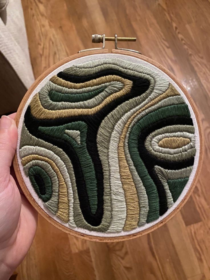 someone is holding up a small embroidery project on a wooden surface with wood flooring in the background