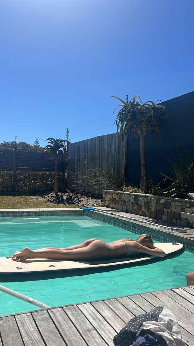 a woman laying on top of a surfboard in a swimming pool next to a wooden deck