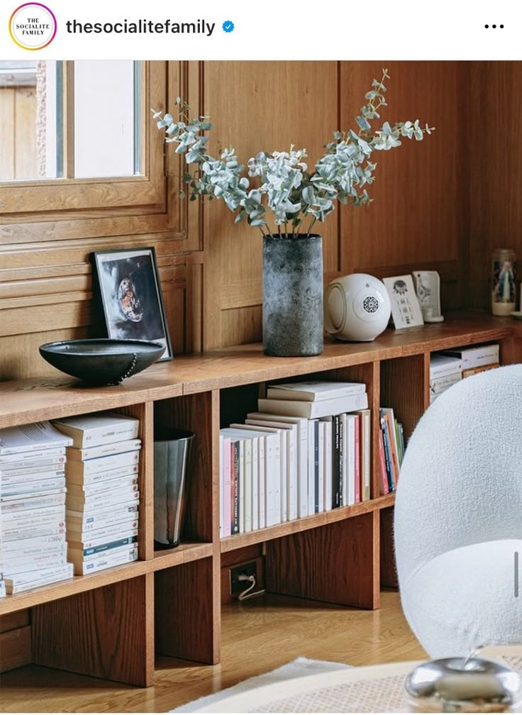 a vase with flowers on top of a wooden shelf next to a chair and bookshelf