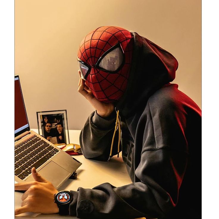 a person sitting at a desk with a laptop and spider man mask on their face