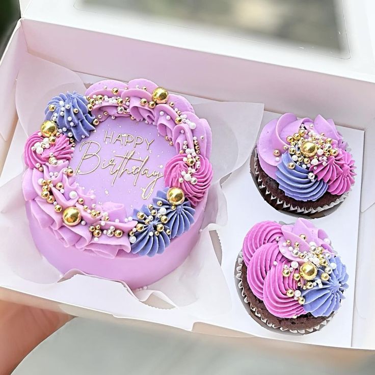 two cupcakes with purple frosting and gold decorations in a pink gift box