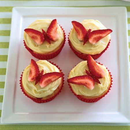 four cupcakes with white frosting and fresh strawberries on top are arranged in a square plate