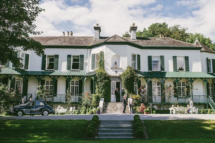 a large white house with green shutters on the front and second story, surrounded by lush greenery