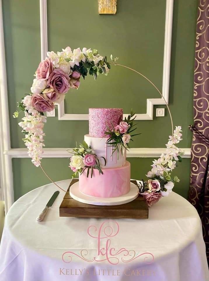 a pink and white wedding cake on a table with greenery around the top, surrounded by flowers